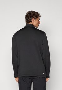 Chaqueta de pulóver negra con cuello alto y mangas largas. Presenta una textura suave, un diseño minimalista y un logo en el lado inferior.