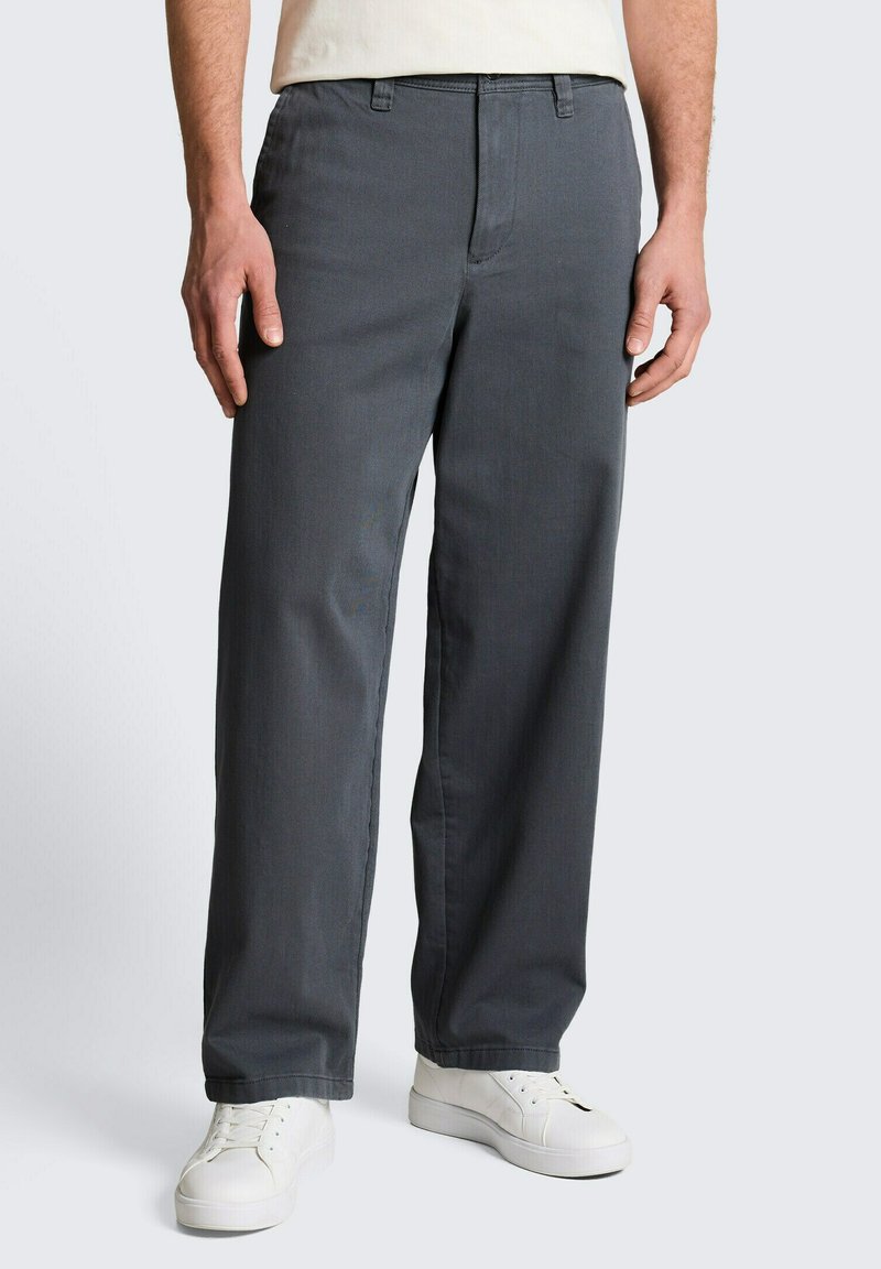 Mann trägt dunkelgraue, gerade geschnittene Hose und weiße Turnschuhe, steht vor einfachem, hellem Hintergrund.