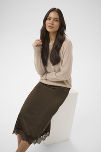 Femme aux longs cheveux foncés portant un pull beige et une jupe en satin marron avec bordure en dentelle, assise sur un cube blanc devant un fond uni.