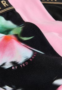 Kukkapainokangasta, jossa on eloisat vaaleanpunaiset, vihreät ja mustat värit. Sileä pinta, jossa kultakirjaimin varustettu logotarra.