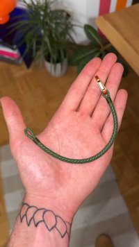 Zeleni pleteni kabel s okruglim krajem i metalnom kopčom, odložen na ruci. Ima teksturiranu površinu. Približna dužina: 15 cm.