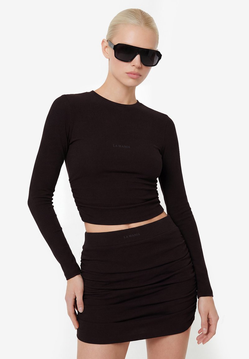 Žena s blond vlasmi, oblečená v čiernom krátkom top s dlhým rukávom a naberanou mini sukňou, doplnená veľkými čiernymi slnečnými okuliarmi.