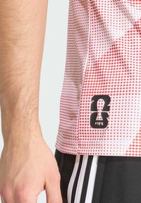 Valkoinen urheilupaita, jossa on punainen timanttikuosi ja musta FIFA-logo helmassa. Sisältää lyhyet hihat ja kevyen, hengittävän kankaan.