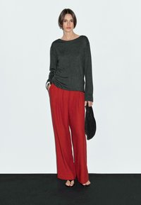 Une femme se tient contre un fond gris clair, portant un pull sombre et un pantalon rouge, tenant un sac noir et affichant une expression neutre.