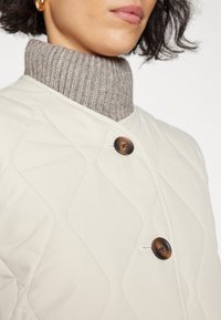 Personne portant une veste matelassée beige avec de grands boutons marron et un pull à col roulé côtelé gris, boucle d'oreille en or visible.