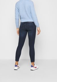 Donkerblauwe skinny jeans met een middelhoog taille, achterzakken en een gladde textuur, gecombineerd met een lichtblauwe gebreide trui en witte sneakers.