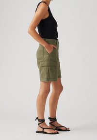 Pantalones cortos cargo verde olivo con dos bolsillos frontales, cinturilla ajustable y dobladillo enrollado, combinados con sandalias negras de tiras y una blusa sin mangas.