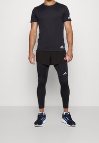 Svart sportt-shirt med Adidas-logotyp, ihop med svarta shorts och leggings. Skorna är marinblå med blå ränder och har vita sulor.
