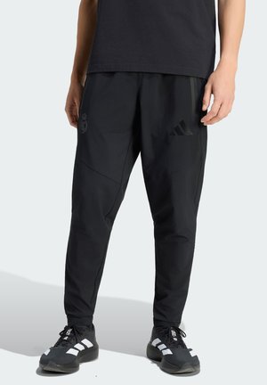 Inimene, kes kannab musta sportlikku pükst, millel on Real Madridi ja Adidase logo, sobitatud mustade ja valgete Adidase tossudega helel taustal.