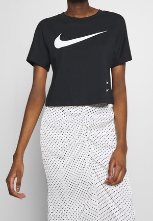 Personne portant un t-shirt Nike court noir avec un grand logo swoosh blanc et une jupe blanche froncée à pois noirs.