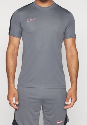 Homme portant un t-shirt de sport gris Nike à manches courtes et un short assorti avec des logos roses, debout, bras détendus le long du corps.