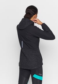 Svart idrottsjacka med grå fleecehuva, med en strömlinjeformad design, reflexdetaljer och textilerad tyg. Visar bakvy.