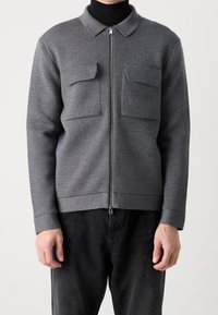 Šedá bunda na zip s límcem, dvěma kapsami na hrudi, žebrovanými manžetami a lemem. Vyrobená z texturované látky, nošená přes černý rolák.