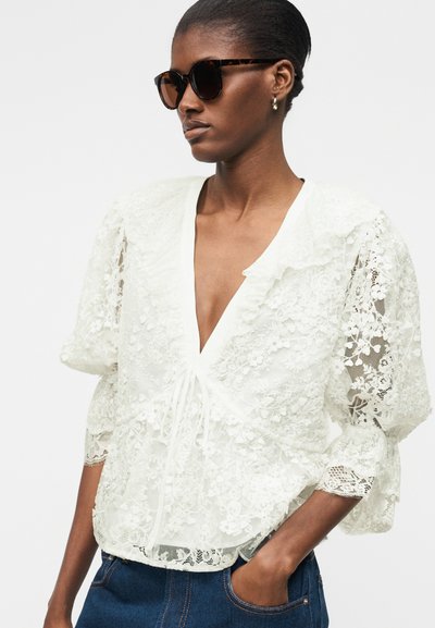 Femme portant des lunettes de soleil oversize en écaille de tortue et un chemisier blanc en dentelle fleurie avec des manches bouffantes et un profond décolleté en V.