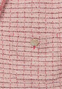 Roze en witte textuurstof met een ruitpatroon, voorzien van een metalen knoop en rode stiksels op een zakgebied.