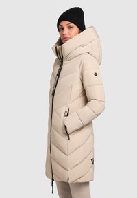 Beige puffer jacket with a high collar, chevron stitching, front zip, and large hood. Paired with a black knitted beanie.