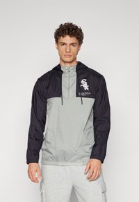 Windbreaker jacket with a gray body and black sleeves, features a hood, drawstring, and logo on the chest, made from lightweight material.