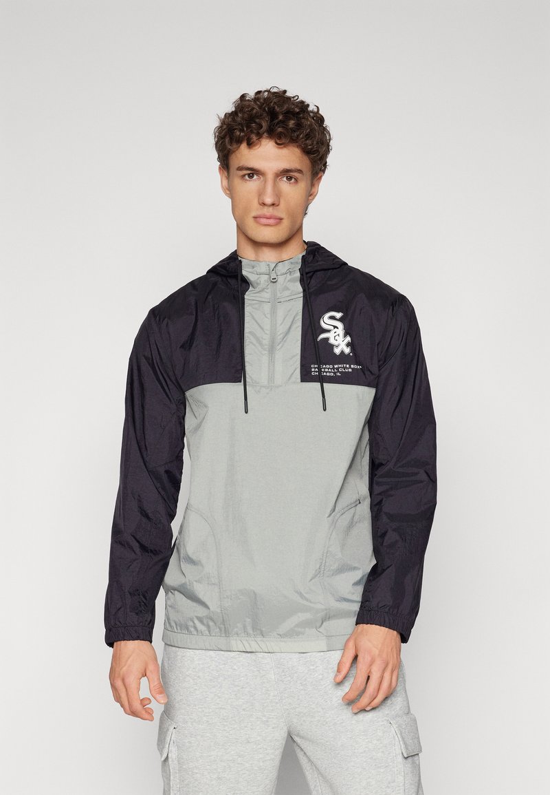 Windbreaker jacket with a gray body and black sleeves, features a hood, drawstring, and logo on the chest, made from lightweight material.