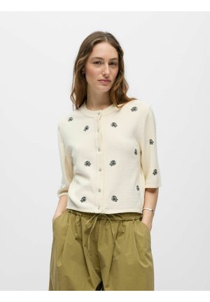 Donna con capelli mossi che indossa un cardigan crema abbottonato con motivi di rose nere e pantaloni larghi verde oliva con coulisse, mani nelle tasche.