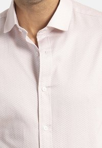 Camisa rosa claro con un sutil patrón geométrico, botón en la parte delantera y un cuello clásico. Textura suave con un diseño ajustado.