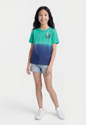 Dziewczyna o długich czarnych włosach, ubrana w zielono-niebieski t-shirt z gradientem, jasne krótkie spodenki jeansowe i białe trampki, uśmiechająca się i stojąca na białym tle.