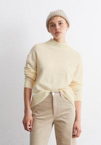 Pulover ușor, de culoare crem, cu gât înalt, mâneci răsucite și tiv cu nervuri, asortat cu pantaloni bej din catifea cu buzunare frontale.