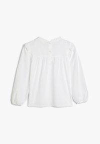 Witte blouse met lange mouwen van gestructureerde stof. Heeft een geplooid lijfje, een halsruffel en een sluiting met knopen op de achterkant met vier knopen.