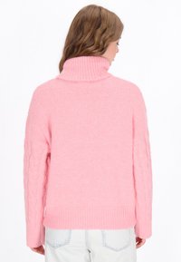 Pulover roz cu guler înalt, fabricat dintr-un material moale și pufoș, cu mâneci tricotate în model cable-knit și tiv zimțat. Vederea din spate arată o croială lejeră.