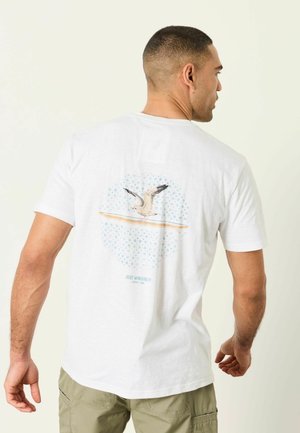 Man wearing white T-shirt with seagull graphic and text "Just Winging It" on the back, paired with light green cargo pants.