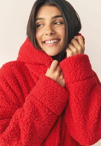 Rote, flauschige Kapuzenjacke mit einer weichen Textur, breiten Ärmeln und einem hohen Kragen. Der Stoff wirkt weich und warm, ideal für kaltes Wetter.