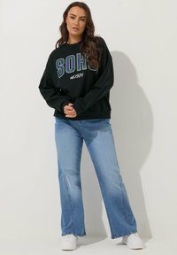 Sudadera negra con el texto "SOHO" y "est. 1978", combinada con unos vaqueros de pierna ancha color azul claro y zapatillas blancas. Mangas largas, corte relajado.