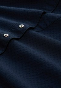 Tessuto di un blu navy con una trama sottile, caratterizzato da due bottoni perlati e una finitura liscia lungo il colletto e la pattina.