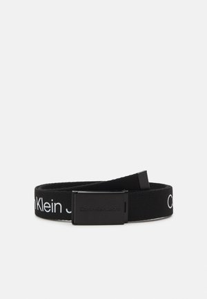 Fekete szövetöv fémcsattal, amelyen fehér "Calvin Klein Jeans" szövegminta található az öv mentén. Mintázott felület.