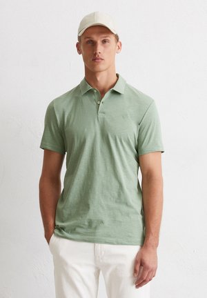 Hellgrünes Poloshirt mit einer weichen Textur, kurzen Ärmeln und einem dreiknöpfigen Ausschnitt. Kombiniert mit weißen Leinenhosen.