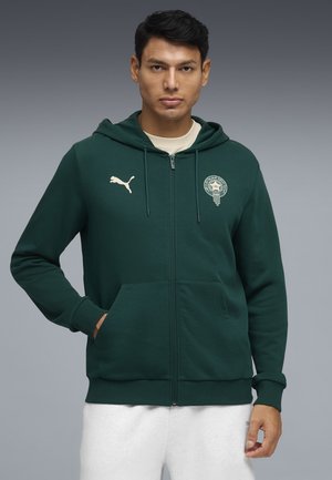 Mann trägt dunkelgrünen Kapuzenpullover mit Reißverschluss, Puma-Logo und rundem Emblem auf der Brust, Hände in den Taschen, vor grauem Hintergrund.