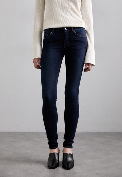 Dunkelblaue Skinny-Jeans mit einer glatten Denim-Oberfläche, einem Reißverschluss und Vordertaschen, kombiniert mit einem cremefarbenen gerippten Pullover und schwarzen Mules.