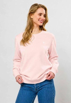 Jeune femme aux longs cheveux blonds souriante, portant un sweat-shirt rose clair et un jean bleu, debout devant un fond uni.