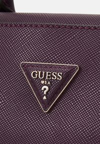 Surface en cuir texturé violet avec un logo triangulaire en métal Guess USA présentant un point d'interrogation au centre.