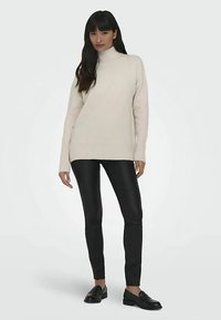 Pull à col roulé crème, poignets et ourlet côtelés, associé à un pantalon noir ajusté et des mocassins noirs. Palette de couleurs simple, texturée et neutre.