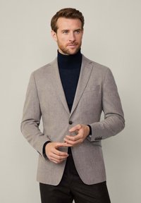 Ljusgrå blazer med texturerad yta, enkel knäppning, slagkrage och bröstficka. Bärs över en marinblå polotröja.