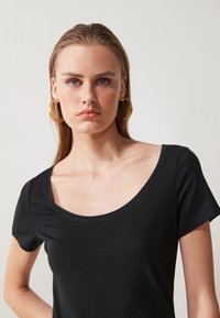 Svart t-skjorte med korte ermer og rund hals, laget av mykt, glatt stoff. Modellen har på seg store, gyldne hooopøreringer. Nøytral bakgrunn.