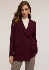 Bordeaux dubbelgebreide blazer met geknipte revers, zes zwarte knopen en voorzakken, gecombineerd met een crèmekleurige top en houndstooth broek.