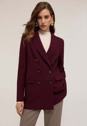 Blazer bordeaux doppiopetto con rever a punta, sei bottoni neri e tasche frontali, abbinato a una maglietta crema e pantaloni houndstooth.