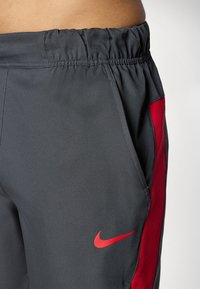 Grå træningsbukser lavet af glat stof, med et rødt sidelæne og et lille rødt Nike-logo. Elastisk talje med samlet tekstur.