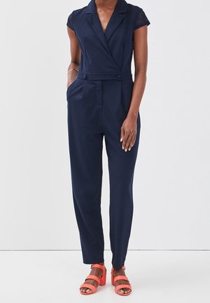 Femme portant une combinaison sans manches bleu marine avec une taille ceinturée et des sandales à talons blocs corail, debout devant un fond uni.