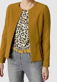 Femme portant une veste jaune moutarde sur un haut à motifs noir, blanc et moutarde, associée à un jean gris clair et une ceinture moutarde.