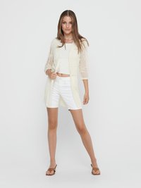 Young woman wearing white knit cardigan, white crop top, white shorts, and brown sandals, posing against plain white background.