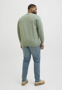 Grüner Baumwollpullover mit Rundhalsausschnitt und langen Ärmeln, kombiniert mit hellblauen Jeans und braunen Stiefeln. Schlichte, lässige Passform.