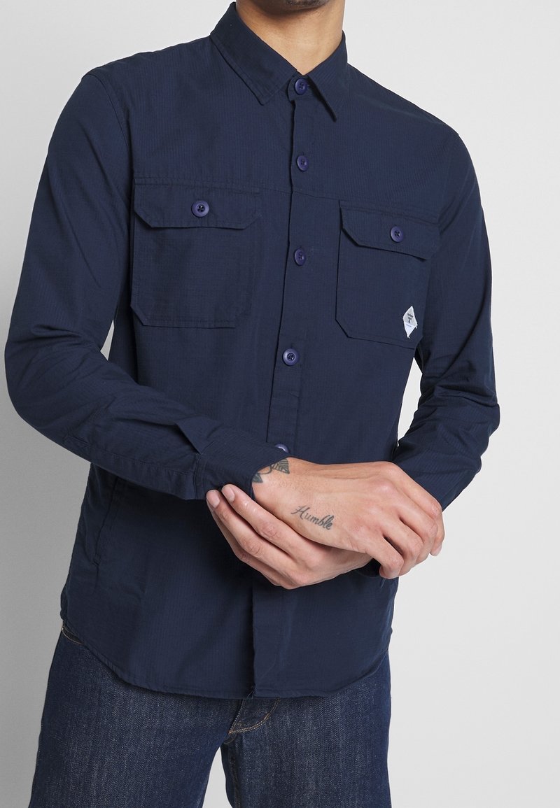 Camicetta a maniche lunghe blu navy realizzata in tessuto testurizzato, con due tasche sul petto, chiusura a bottoni e dettagli viola sui bottoni.