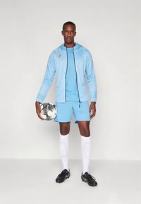 Svetlomodrá športová súprava s čiernymi detailmi, doplnená svetlomodrými šortkami. Postava drží čierno-bielu futbalovú loptu, má na sebe čierne kopačky a biele ponožky.